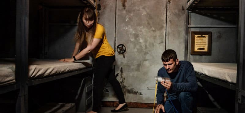 Woman leaning on bed and man kneeling on floor looking for clues